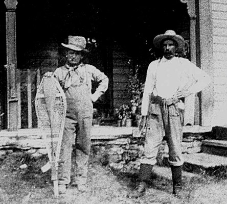 an old image of 2 men standing in front of a house with snowshoes